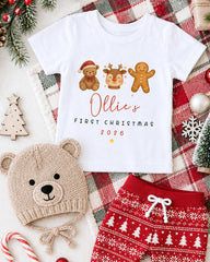 A white t-shirt with a graphic design of three Christmas-themed characters - a teddy bear, a gingerbread man, and a reindeer - along with the text "Ollie's FIRST CHRISTMAS 2006". The t-shirt is displayed on a plaid blanket, and there are other Christmas-themed items such as a knitted hat, a pair of red shorts, and a small Christmas tree in the background.