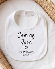 A white bib with the text "Coming Soon" and "Baby Davis 2026" printed on it, placed on a white cloth.