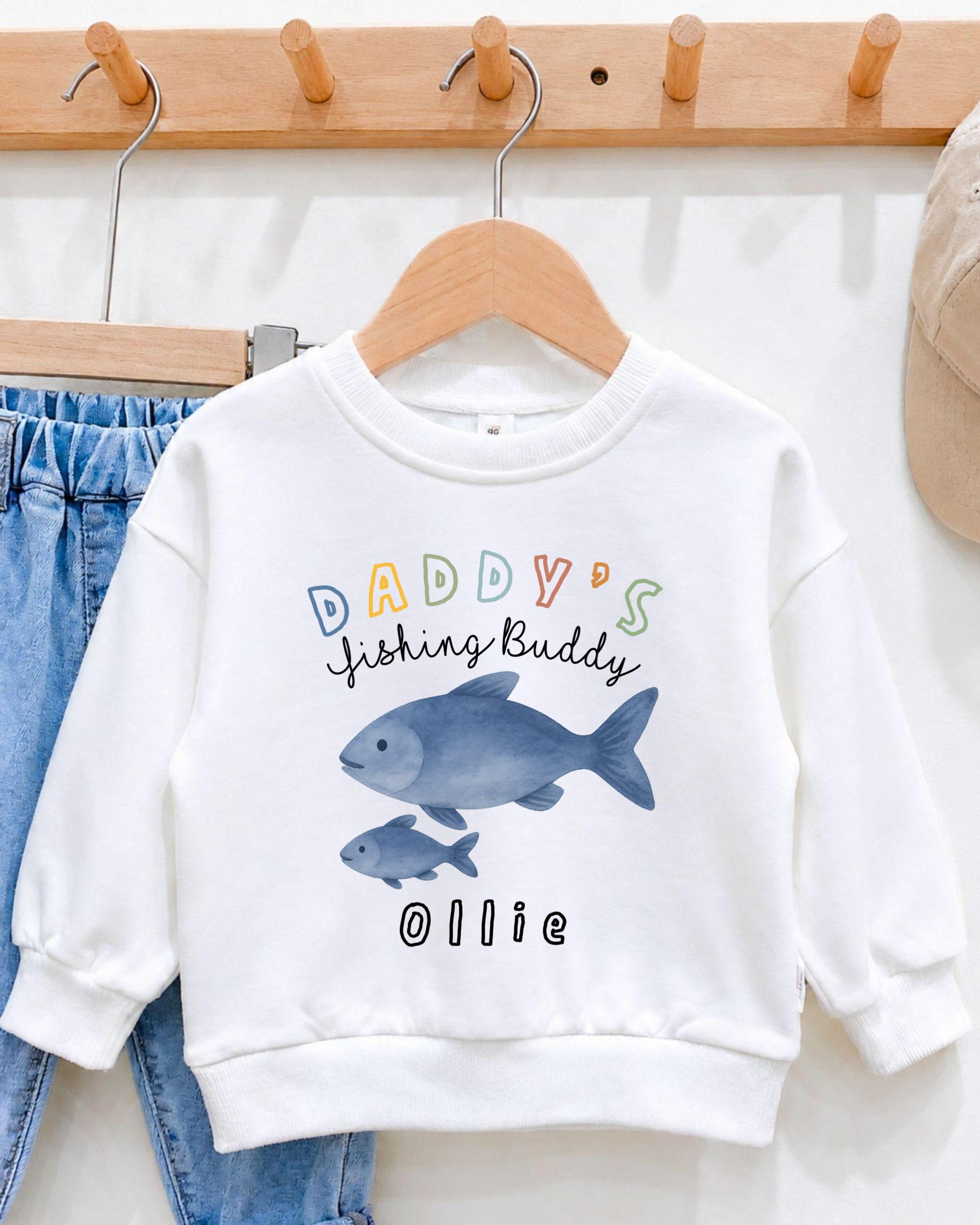 A white sweatshirt with a colorful fish graphic and text that says "DADDY'S Fishing Buddy OLLIE" hanging on a wooden hanger.