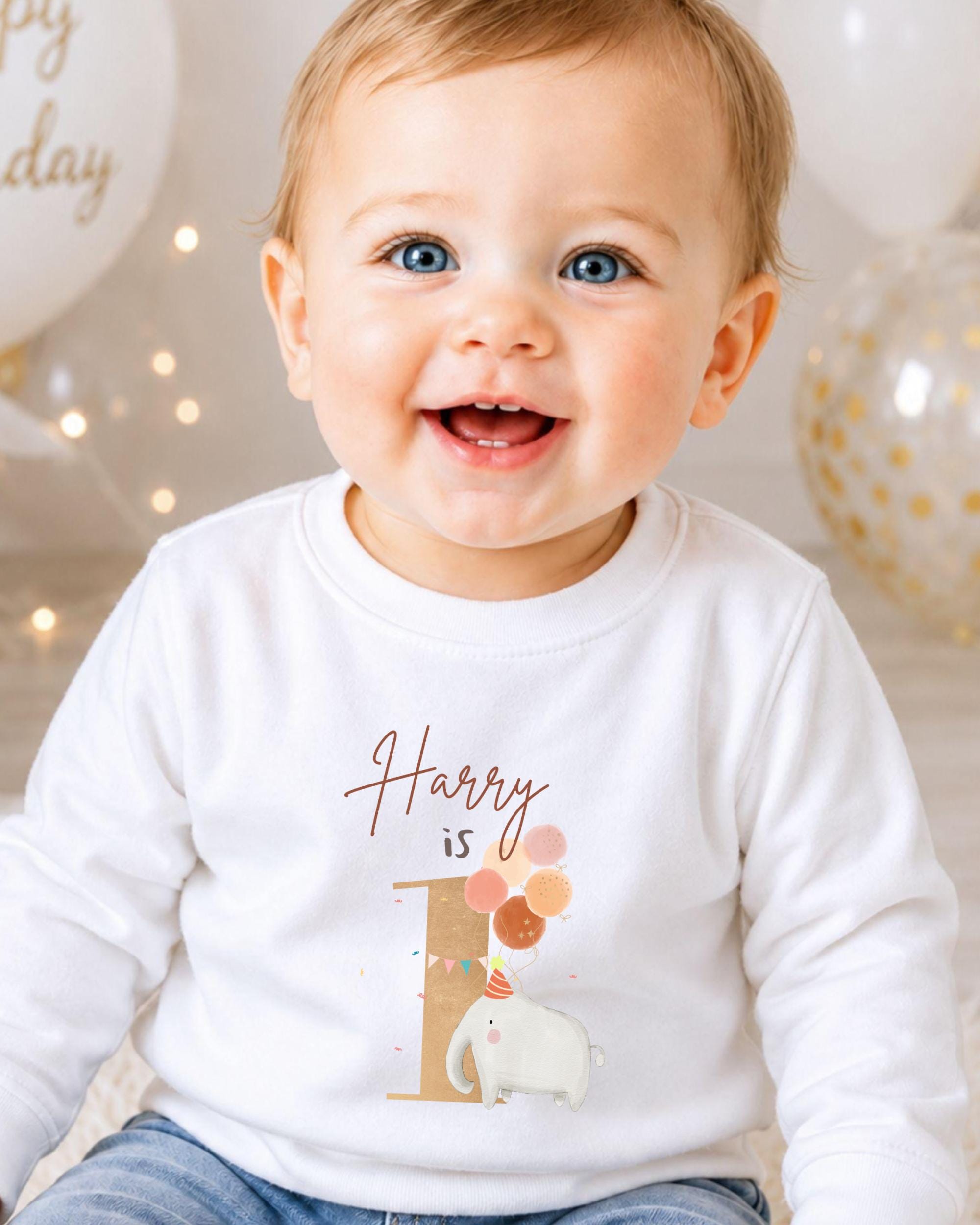 A smiling baby boy wearing a white shirt with a birthday-themed design.