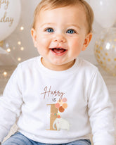 A smiling baby boy wearing a white shirt with a birthday-themed design.