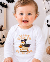 A smiling baby wearing a white onesie with a cartoon goose and the text "Spooky Goose on the Loose" is sitting in front of Halloween decorations.