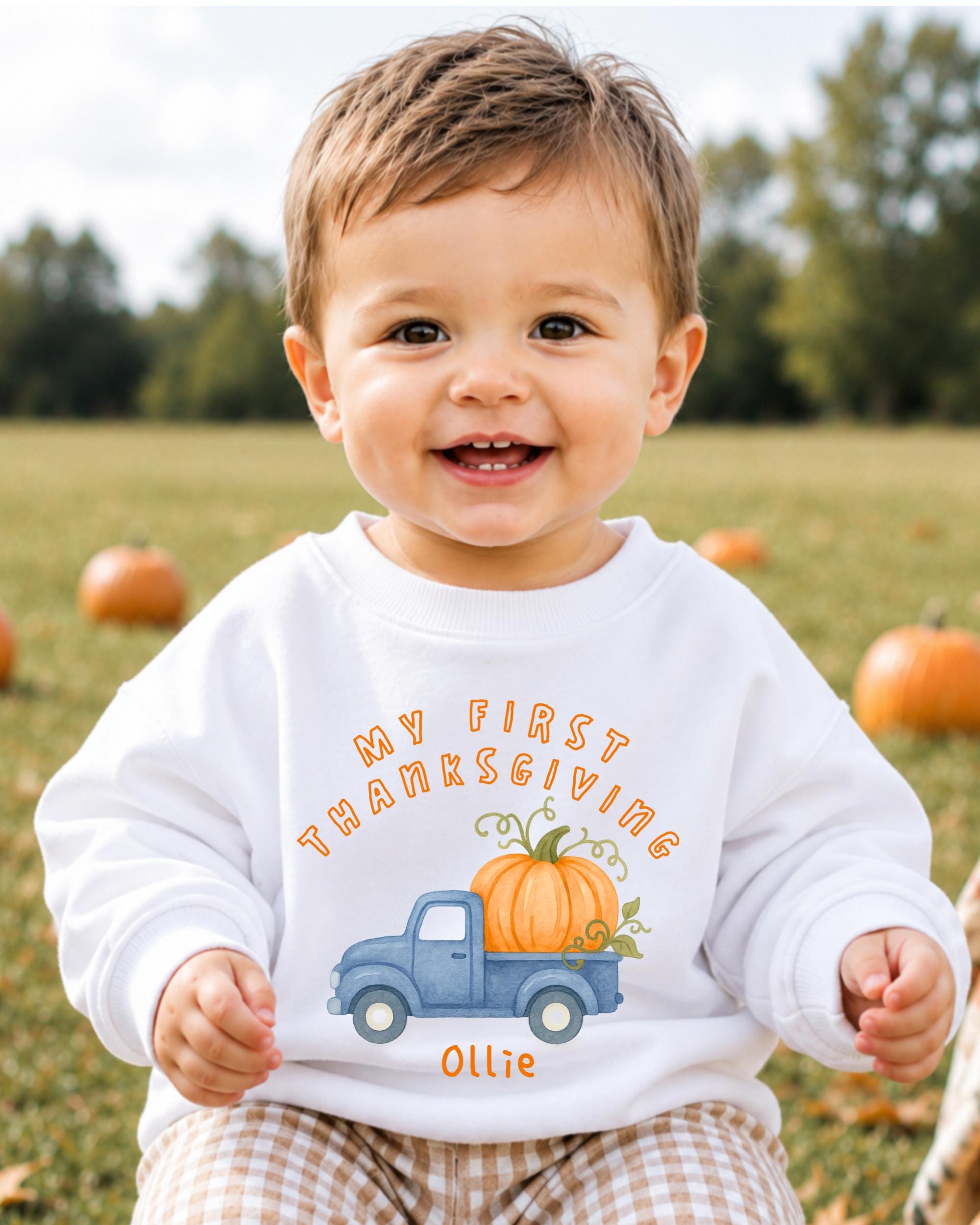 A smiling baby boy wearing a white sweatshirt with a pumpkin and a truck graphic, sitting in a field with pumpkins in the background.
