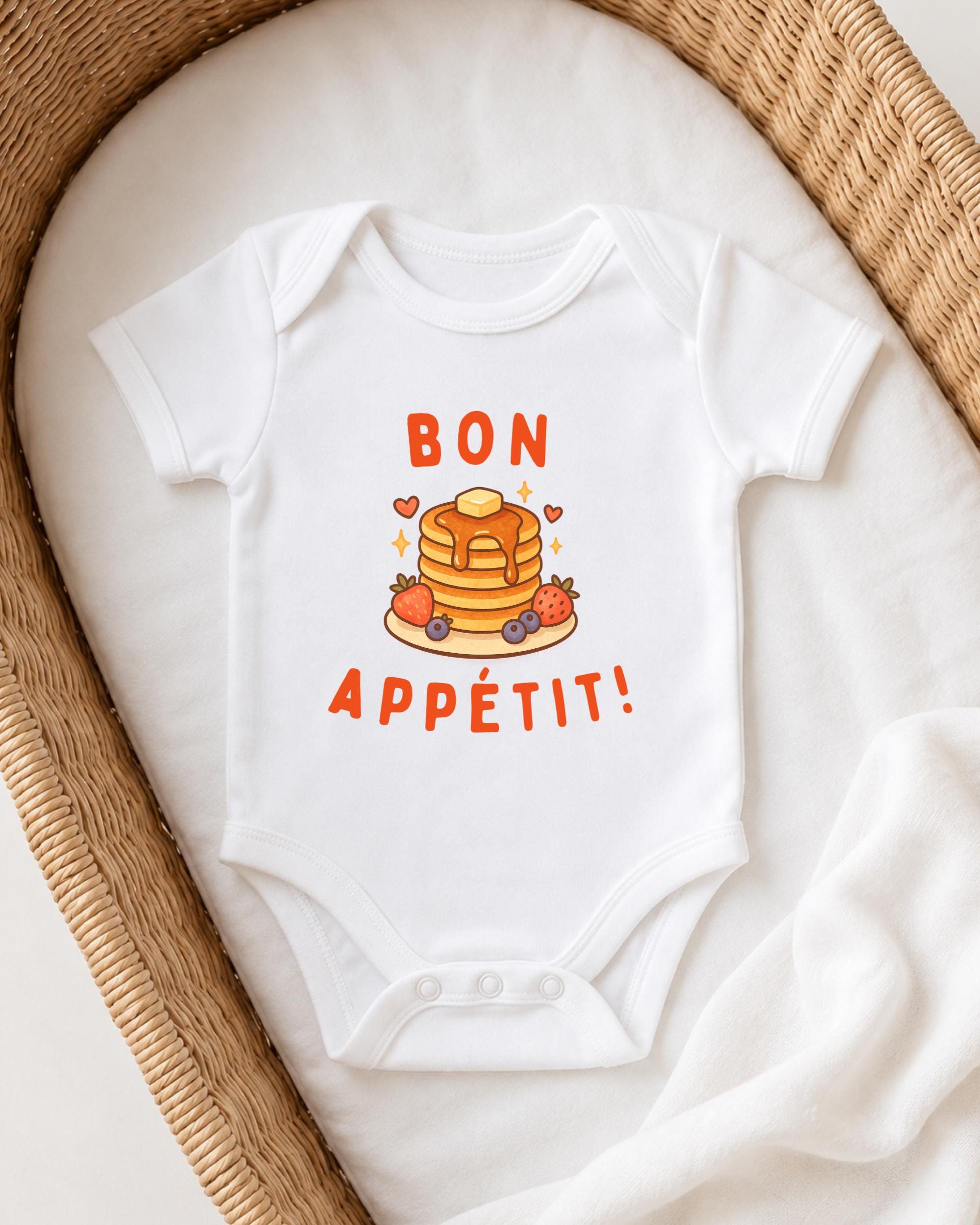 A white onesie with a pancake and fruit design, along with the text "BON APETIT!" printed on it, is displayed on a white blanket.