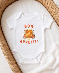 A white onesie with a pancake and fruit design, along with the text "BON APETIT!" printed on it, is displayed on a white blanket.