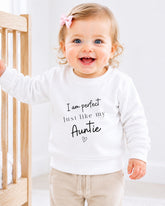A smiling baby girl wearing a white sweatshirt with the text "I am perfect just like my Auntie" printed on it.