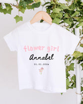 A white t-shirt with pink text and a small floral design hangs on a wooden hanger, surrounded by greenery.
