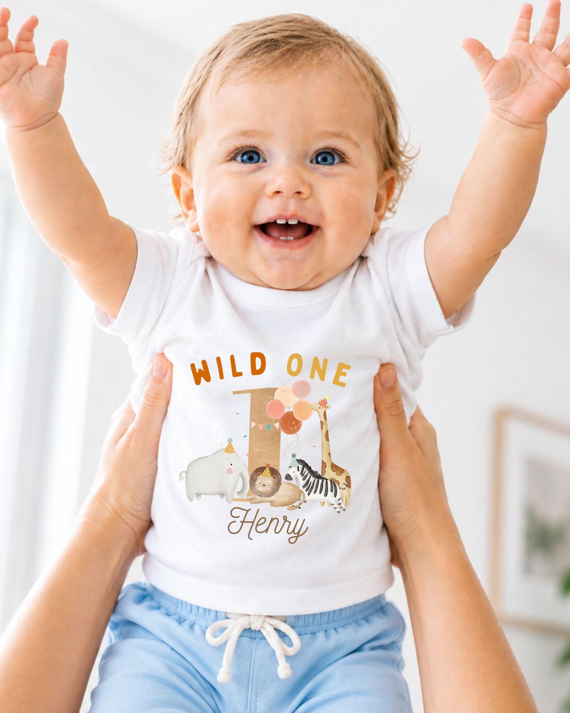 A smiling baby wearing a white t-shirt with a cartoon animal design and the text "Wild One Henry" is being held up by two people.