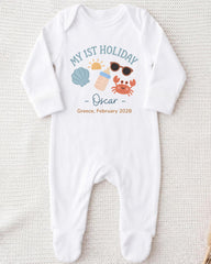 A white onesie with a graphic design on it, featuring icons of a sun, sunglasses, a shell, and a crab, along with the text "My 1st Holiday Oscar - Greece - February 2028".
