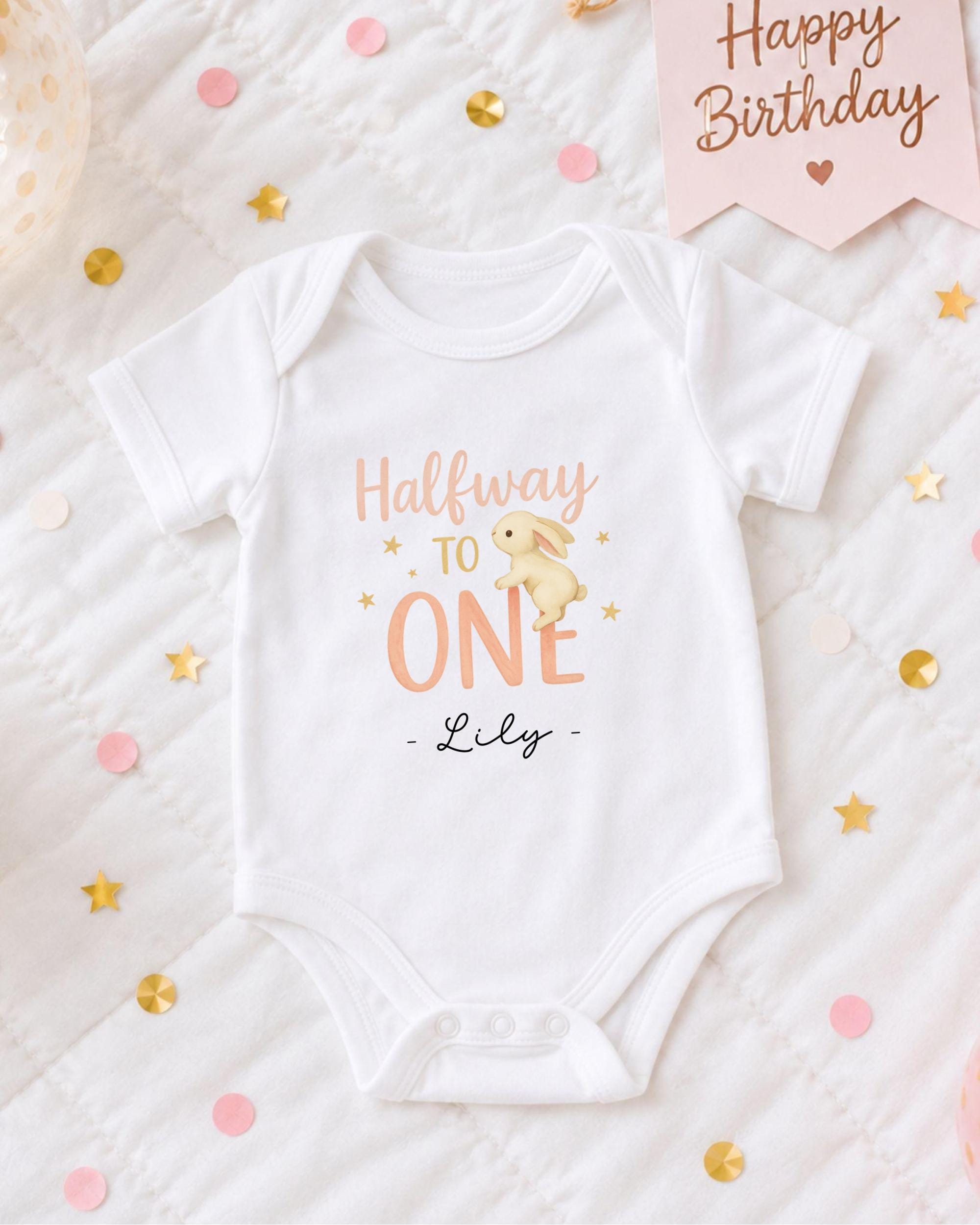 A white onesie with the text "Halfway to one" printed on it, along with a small bunny illustration. The onesie is placed on a bed with confetti and a "Happy Birthday" sign.