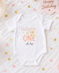 A white onesie with the text "Halfway to one" printed on it, along with a small bunny illustration. The onesie is placed on a bed with confetti and a "Happy Birthday" sign.