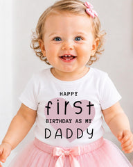 A smiling baby girl wearing a white t-shirt with text and a pink bow, standing in front of a white background.
