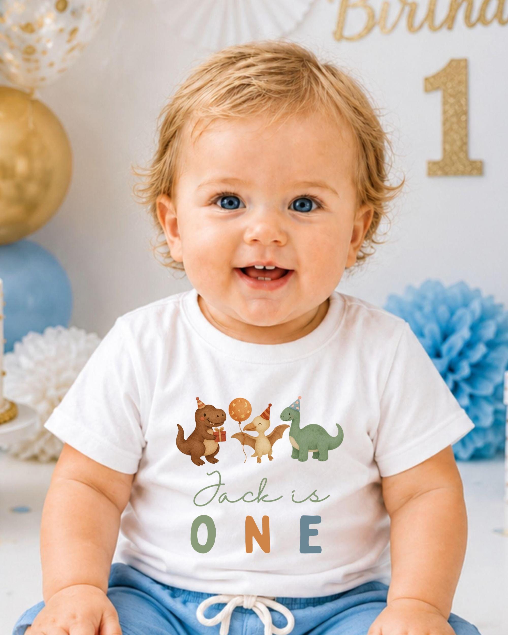 A smiling baby boy wearing a white t-shirt with dinosaur illustrations and the text "Jack is one" is sitting in front of a birthday party backdrop.