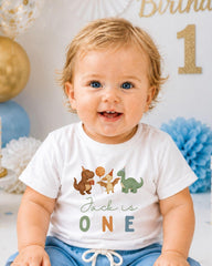A smiling baby boy wearing a white t-shirt with dinosaur illustrations and the text "Jack is one" is sitting in front of a birthday party backdrop.