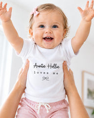 A smiling baby girl wearing a white t-shirt with the text "Auntie Hollie loves me" is being held up by someone's arms.