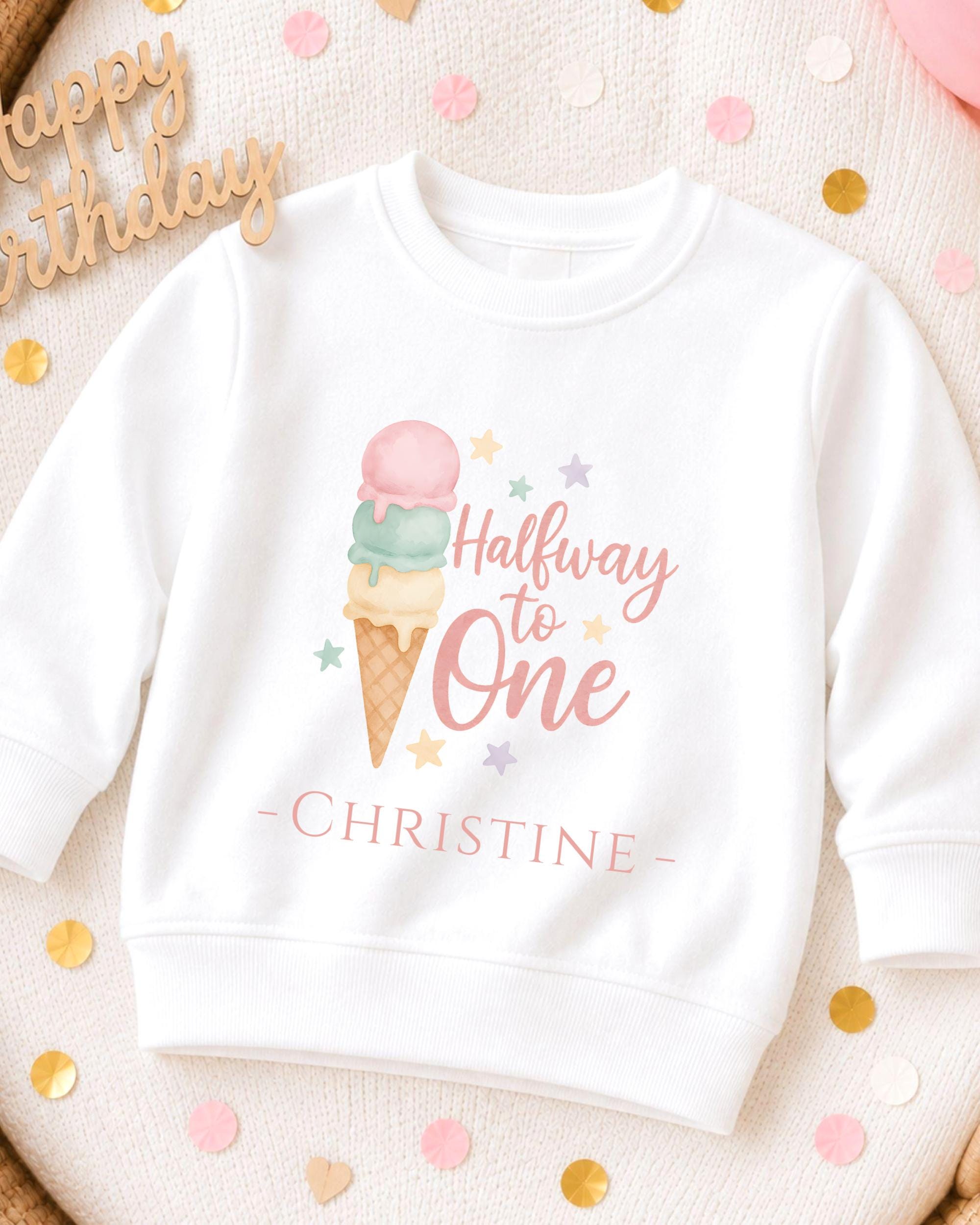 A white sweatshirt with a colorful graphic of an ice cream cone and the text "Halfway to One" printed on it, along with the name "Christine" below. The sweatshirt is placed on a surface with scattered confetti.