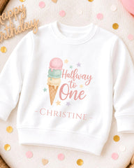 A white sweatshirt with a colorful graphic of an ice cream cone and the text "Halfway to One" printed on it, along with the name "Christine" below. The sweatshirt is placed on a surface with scattered confetti.