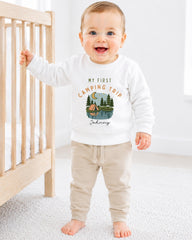 A smiling baby boy wearing a white sweatshirt with a camping trip design and the text "My first camping trip" is standing next to a wooden crib.