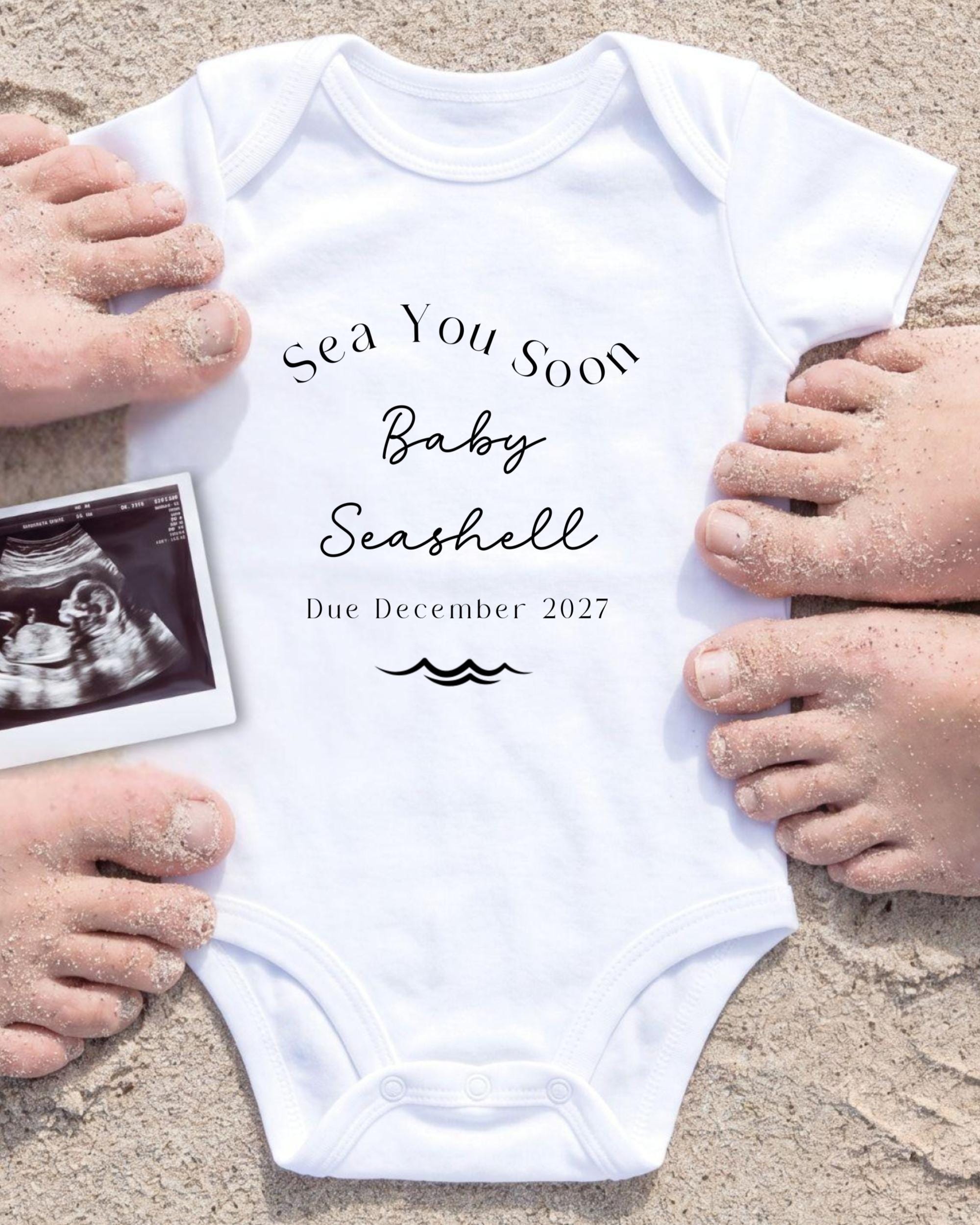 a white onesie with text and an image of an Ultrasound scan of a baby's belly. The text on the onesie reads "Sea You Soon Baby Seashell Due December 2027".