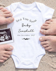 a white onesie with text and an image of an Ultrasound scan of a baby's belly. The text on the onesie reads "Sea You Soon Baby Seashell Due December 2027".