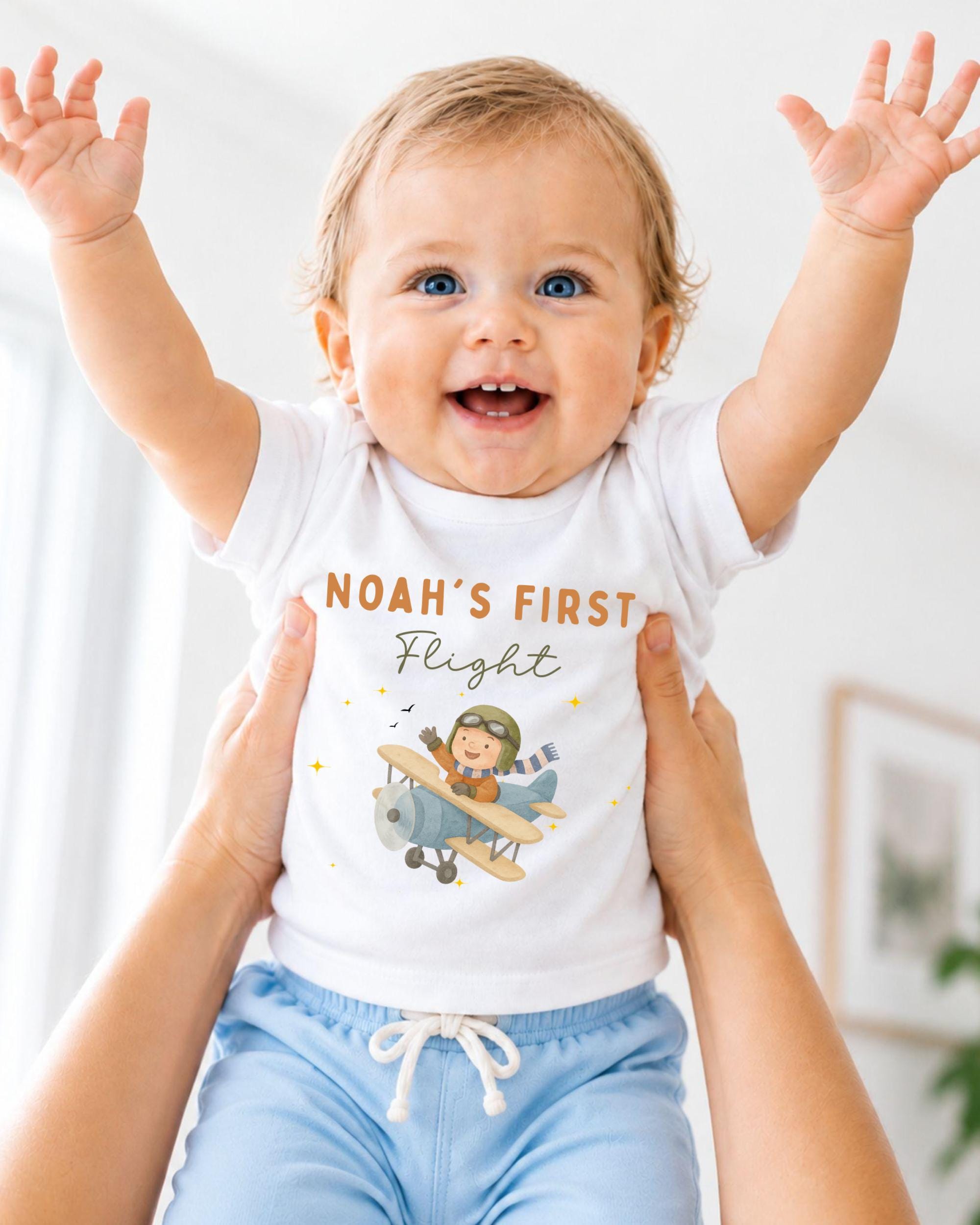 A smiling baby wearing a white t-shirt with a cartoon airplane and the text "Noah's first flight" is being held up by two people.