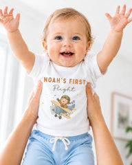 A smiling baby wearing a white t-shirt with a cartoon airplane and the text "Noah's first flight" is being held up by two people.