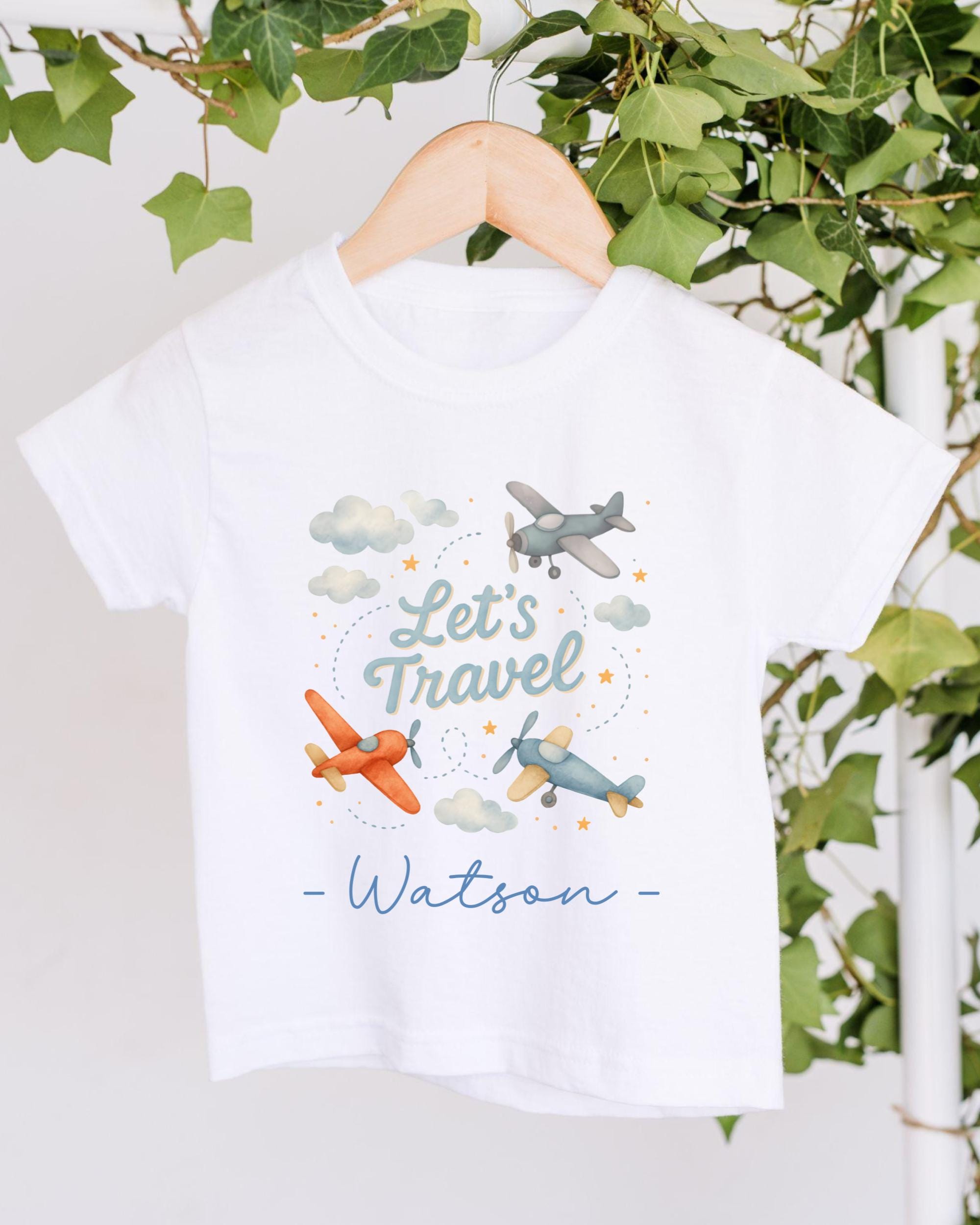 A white t-shirt with a graphic design featuring two airplanes and the text "Let's Travel" along with a cloud and star pattern.