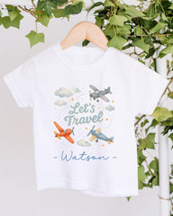 A white t-shirt with a graphic design featuring two airplanes and the text "Let's Travel" along with a cloud and star pattern.