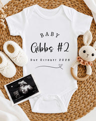a baby announcement, including a white onesie with the text "BABY Gibbs #2 Due October 2026", a crocheted bunny toy, and a black and white photograph of an Ultrasound image.