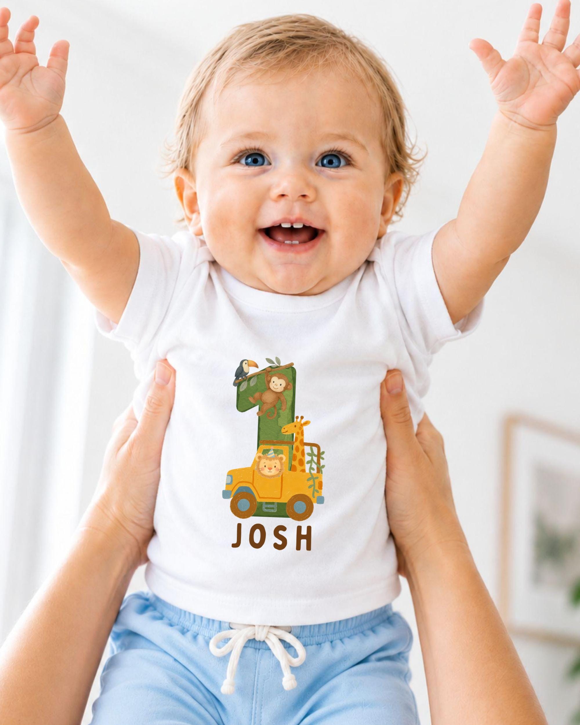 A smiling baby wearing a white t-shirt with a cartoon character and the name " Josh" printed on it, being held up by someone's arms.