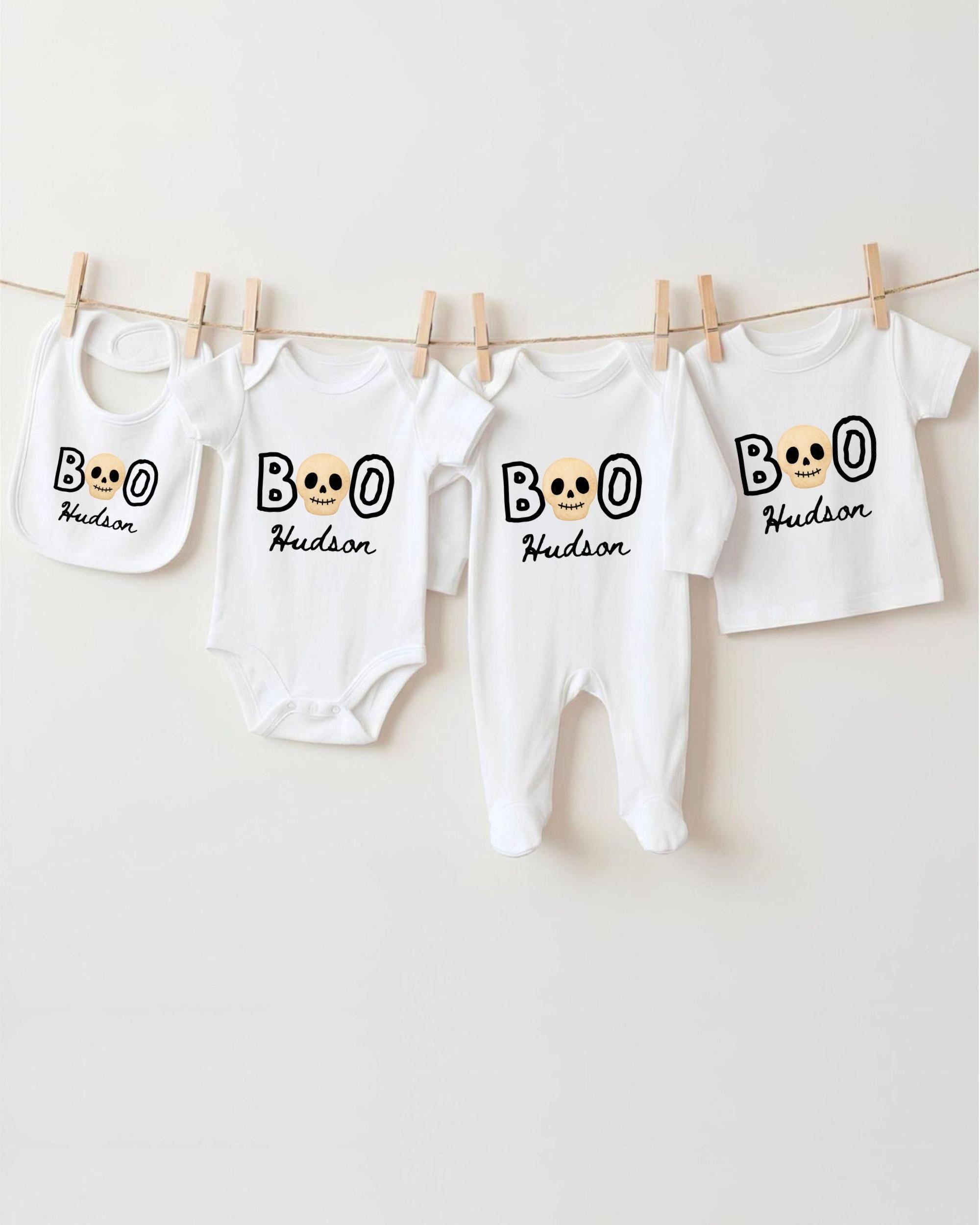 A collection of white baby clothes, including onesies and bibs, with the name "Hudson" printed on them. The clothes are hanging on a clothesline, with the name "BOO" printed on them.