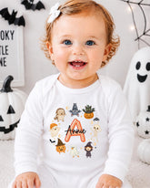 A smiling baby girl wearing a white onesie with a personalized name on it, surrounded by Halloween decorations.