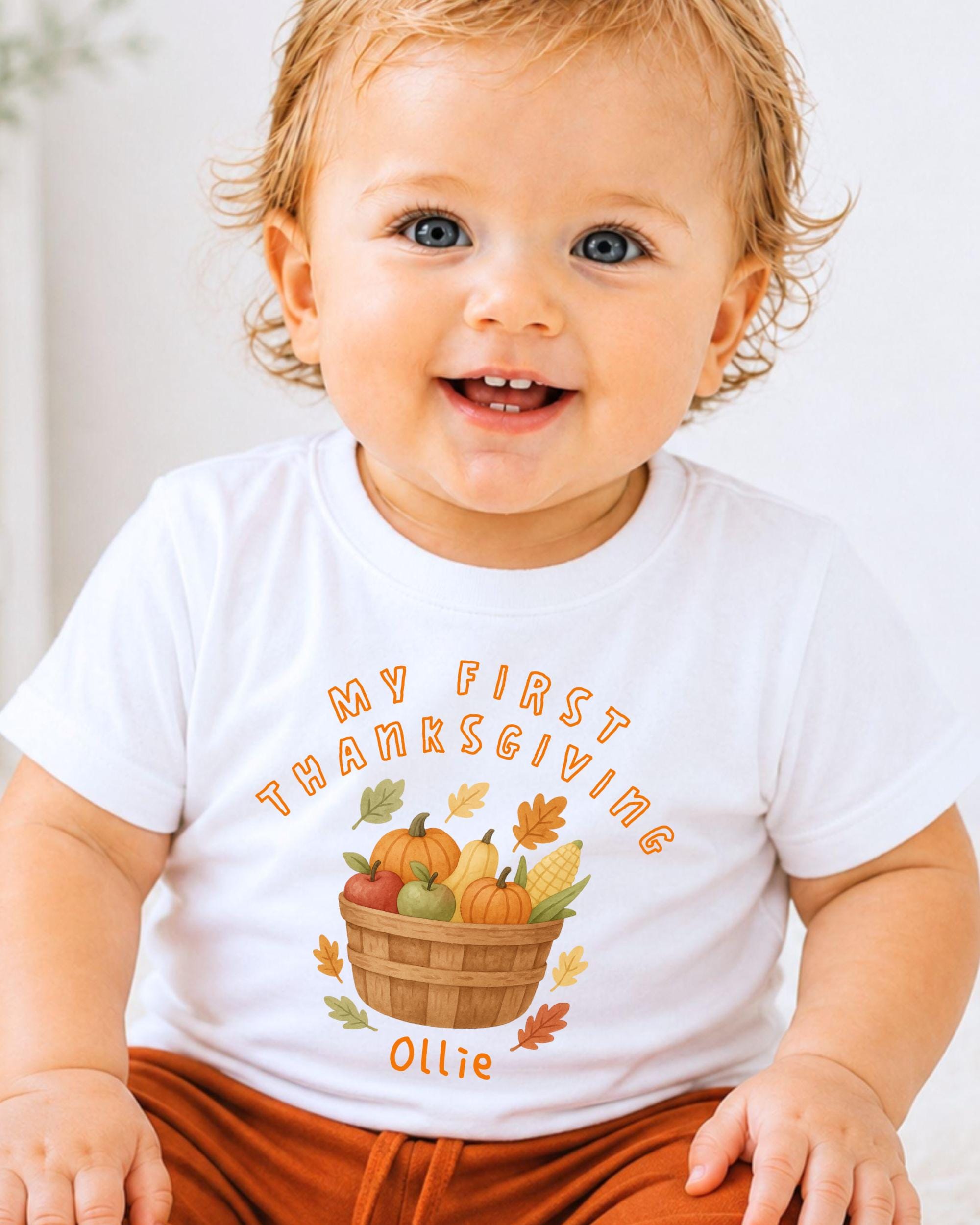 A smiling baby wearing a white t-shirt with a graphic of a basket of fall fruits and vegetables, along with the text "My first Thanksgiving Ollie".