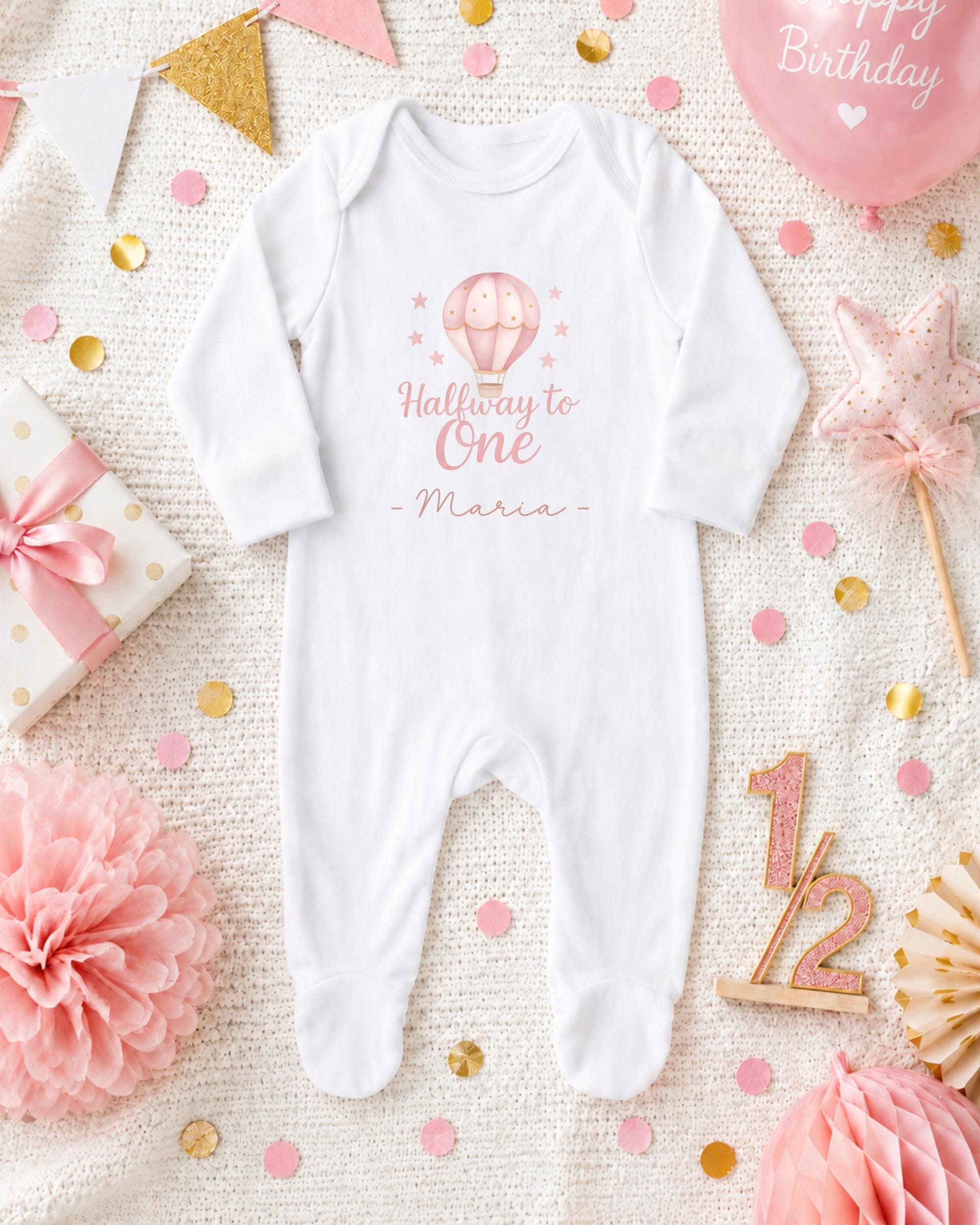 A white onesie with a balloon and stars on it, surrounded by pink and gold confetti, a gift box, and a star-shaped decoration.