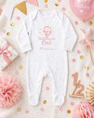 A white onesie with a balloon and stars on it, surrounded by pink and gold confetti, a gift box, and a star-shaped decoration.