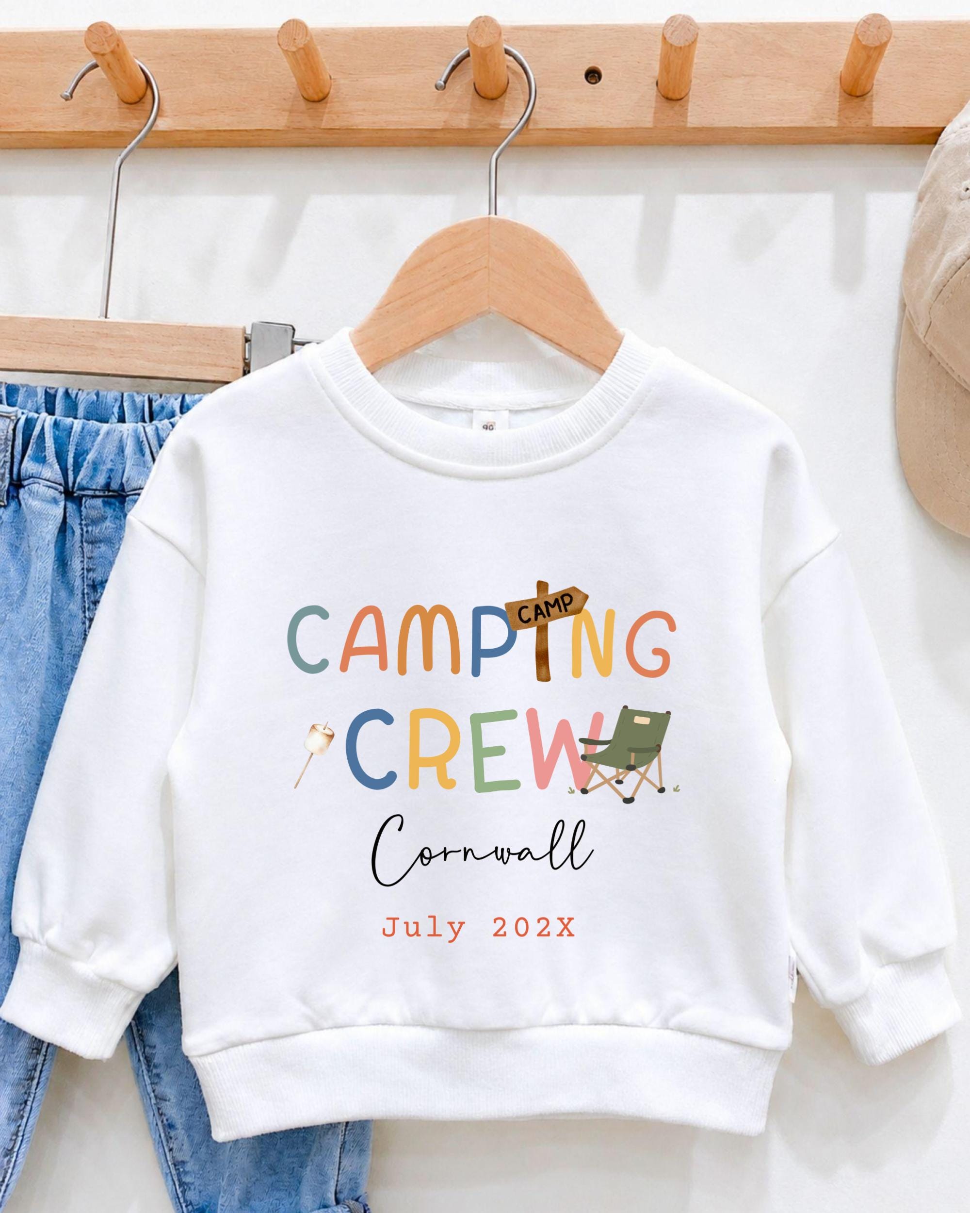 A white sweatshirt with the text "CAMPING CREW Cornwall July 202x" printed on it, along with various camping-related icons and text.