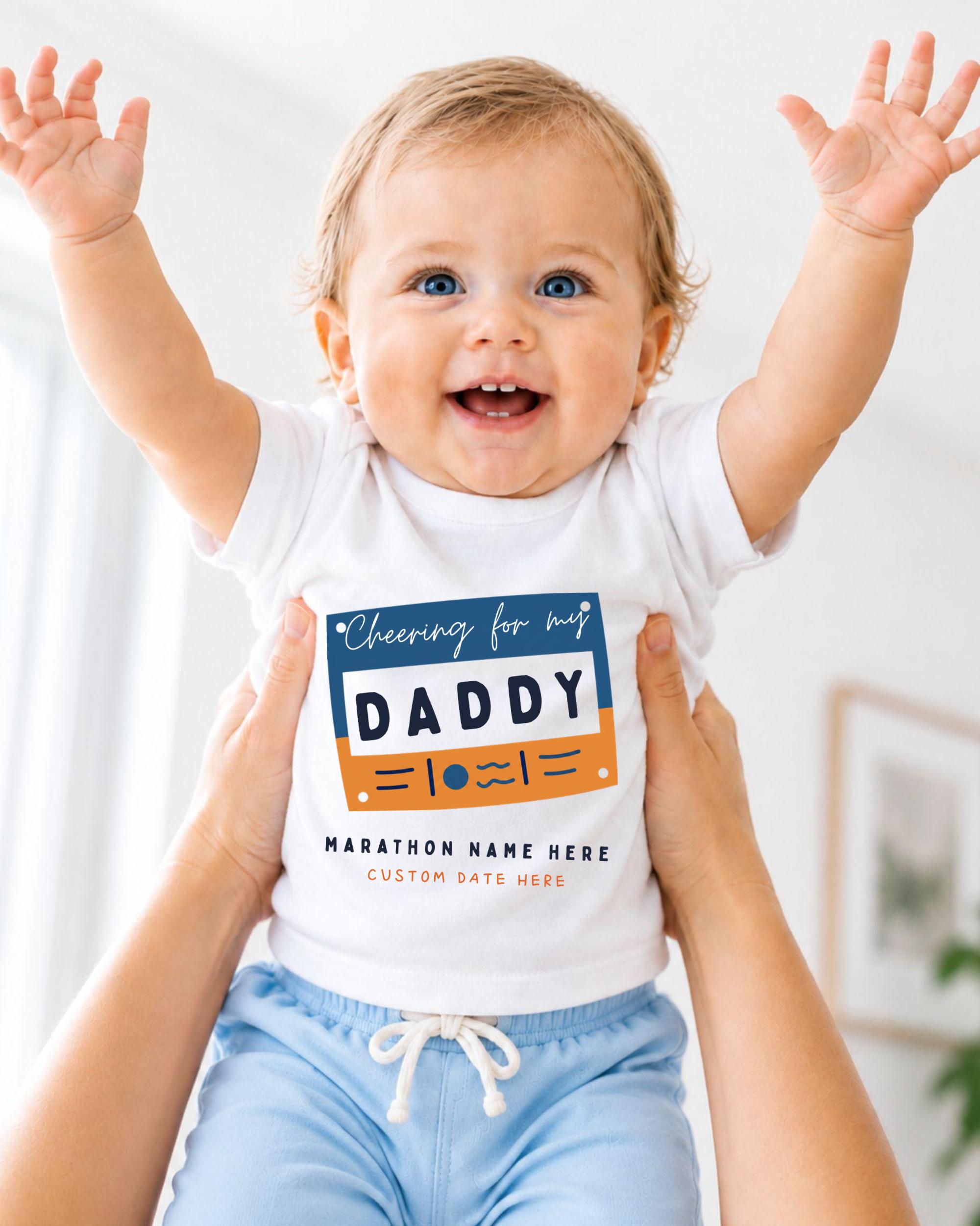 A smiling baby wearing a white shirt with a graphic design and text, being held up by someone's arms.