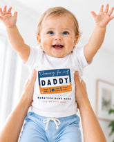 A smiling baby wearing a white shirt with a graphic design and text, being held up by someone's arms.