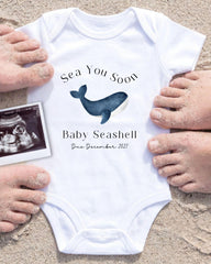 A white baby onesie with a blue whale graphic and text, surrounded by sand and a black and white photograph of a baby's feet.