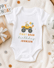 A white onesie with a yellow truck and colorful balloons, along with a "Happy Birthday" sign and a gift box, all arranged on a white blanket with confetti.