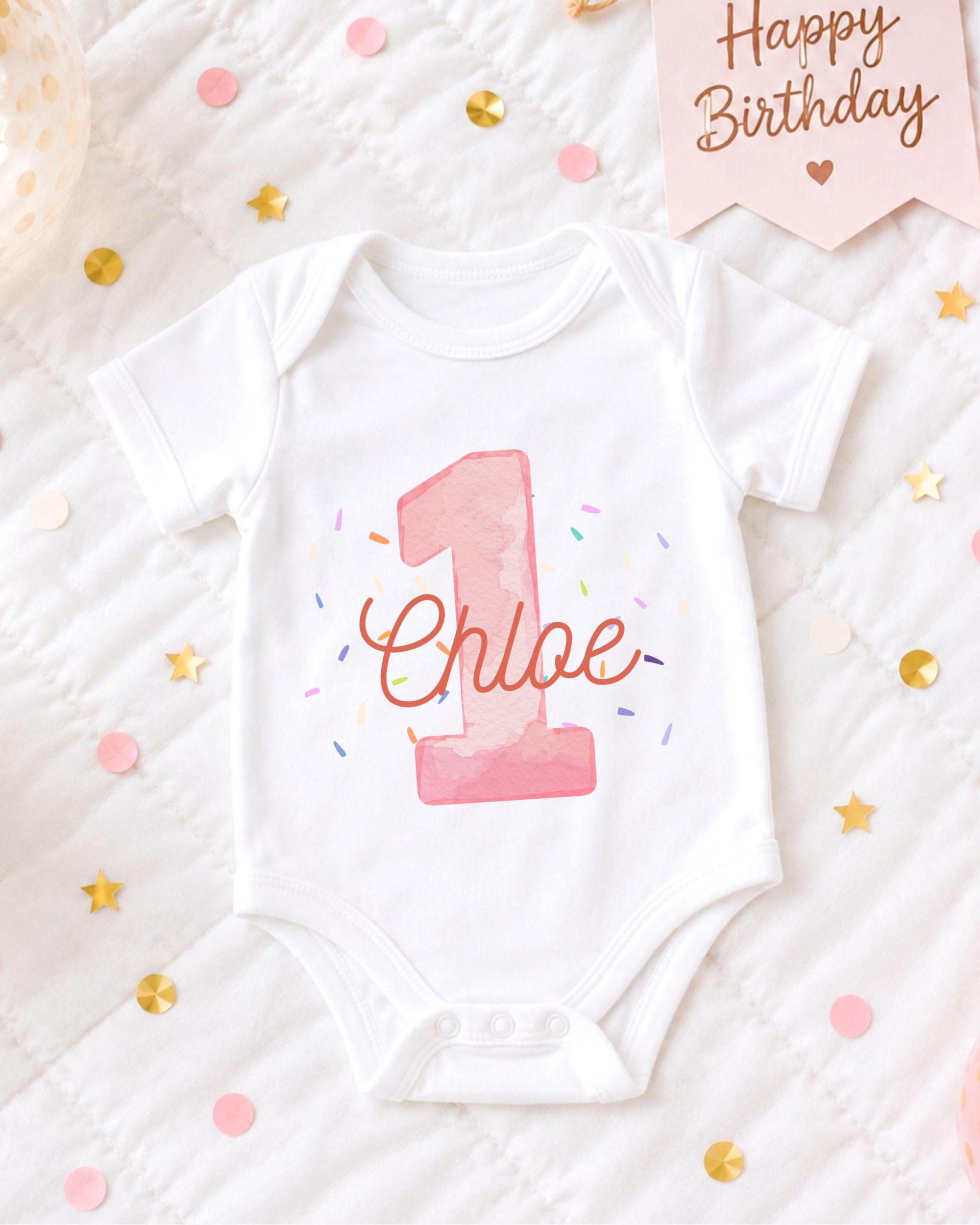 A white onesie with the number 1 and the name "Chloe" printed on it, surrounded by confetti and a birthday card.