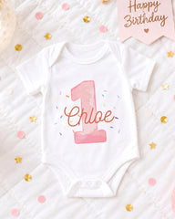 A white onesie with the number 1 and the name "Chloe" printed on it, surrounded by confetti and a birthday card.