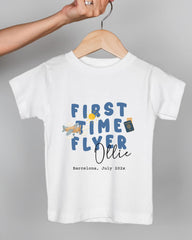 A white t-shirt with a graphic design featuring the text "FIRST TIME FLYER OLLIE" and a small airplane. The text is in blue and orange colors, and the shirt is being held by a person's hand.