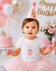 A young girl wearing a white t-shirt with a strawberry design and a pink party hat sits in front of a pink and white birthday cake, surrounded by pink and white balloons and flowers.