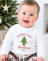 A smiling baby boy wearing a white sweatshirt with a Christmas tree design and text that says "Oliver's first Christmas 2016".