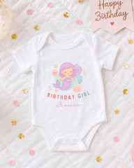 A white onesie with a mermaid design and the text "Birthday Girl Jessica" printed on it, along with confetti and a birthday card.