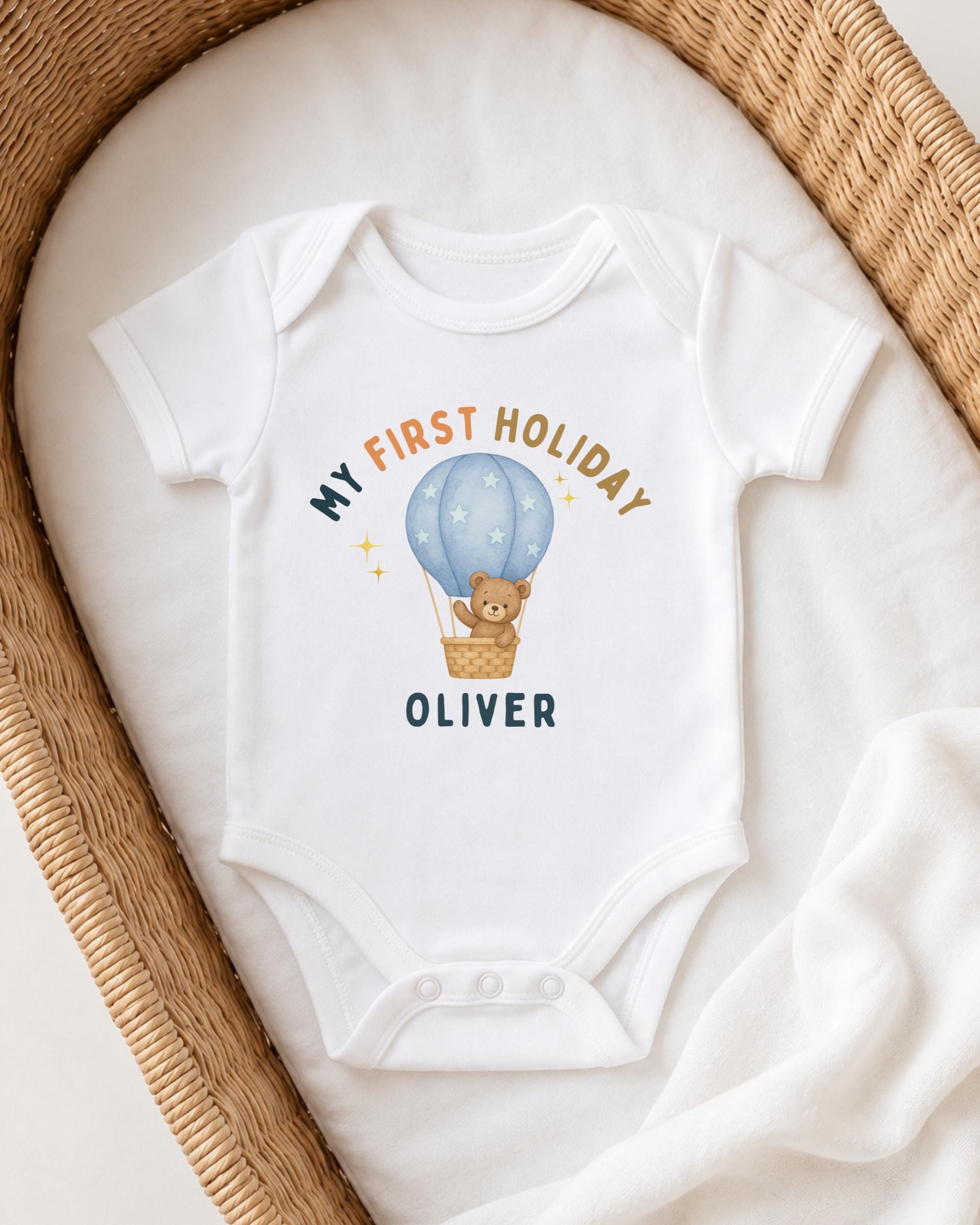 A white onesie with a teddy bear and a hot air balloon graphic printed on it, placed on a white blanket.