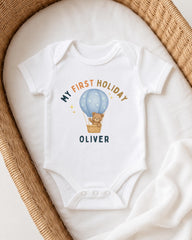 A white onesie with a teddy bear and a hot air balloon graphic printed on it, placed on a white blanket.