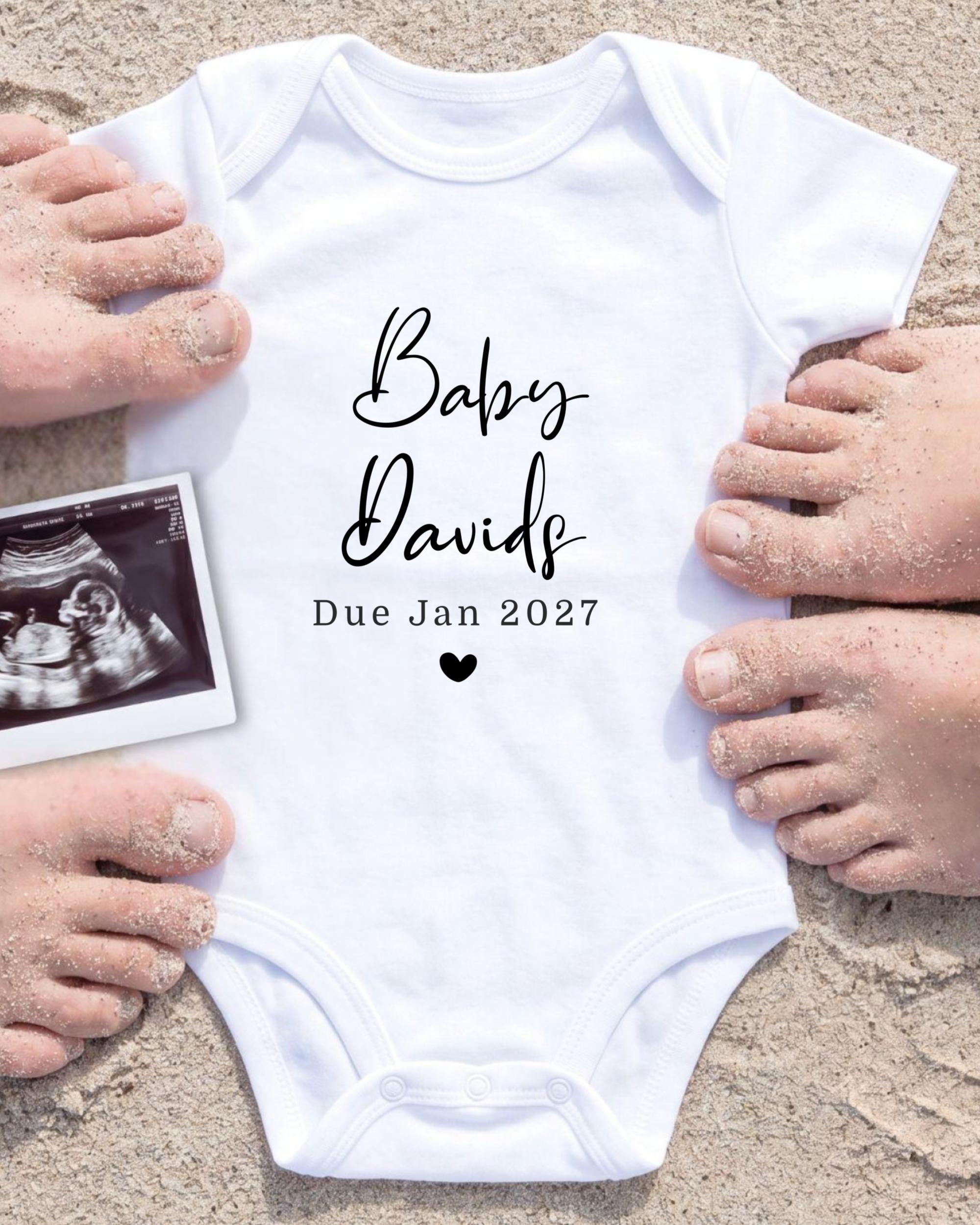 A white baby onesie with the text "Baby Davis Due Jan 2027" printed on it, surrounded by sand and a black and white photograph of a baby's belly.