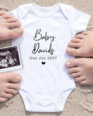 A white baby onesie with the text "Baby Davis Due Jan 2027" printed on it, surrounded by sand and a black and white photograph of a baby's belly.